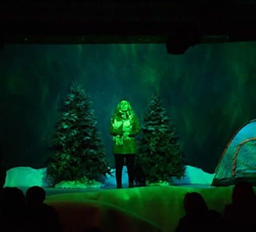 Actress onstage in nighttime scene with tent, pine trees and snow in background.